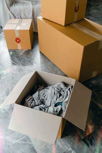 Interior scene showing a home relocation process with several cardboard boxes of varying sizes sealed with beige packing tape, some with red and black hazard or handling symbols, placed on a grey stone floor. One open cardboard box contains loosely packed clothing or fabric. In the background, a partially visible sofa with light-colored upholstery is seen. Additional boxes are stacked near a doorway or entryway, suggesting packing and loading activities typical of furniture transport and house removals. Equipment such as a trolley or straps is not visible. The environment appears to be a residential interior, with natural light illuminating the space, demonstrating typical components involved in packing for a moving service provided by Movers Balham.