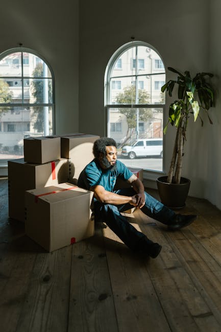 A man with curly hair sits on a wooden floor inside a room with white walls and large arched windows, through which a street with parked cars and modern buildings can be seen. He is wearing a teal T-shirt and dark pants, and is resting beside a stack of cardboard moving boxes, some sealed with red tape. The boxes vary in size and are arranged haphazardly, indicating they are part of a packing process during home relocation. The room is illuminated by natural light coming through the windows, casting shadows on the floor and highlighting the man's contemplative expression. To his right, a tall potted plant with large green leaves adds a touch of greenery to the space. This scene reflects the interior environment during furniture transport and packing activities associated with professional removals by companies such as Movers Balham in the context of house moves, compatible with the Balham High Road removals guide.
