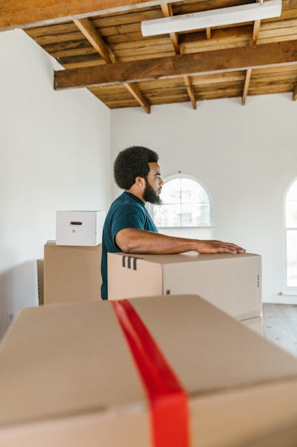 Interior scene showing a home relocation process with several cardboard boxes of varying sizes sealed with beige packing tape, some with red and black hazard or handling symbols, placed on a grey stone floor. One open cardboard box contains loosely packed clothing or fabric. In the background, a partially visible sofa with light-colored upholstery is seen. Additional boxes are stacked near a doorway or entryway, suggesting packing and loading activities typical of furniture transport and house removals. Equipment such as a trolley or straps is not visible. The environment appears to be a residential interior, with natural light illuminating the space, demonstrating typical components involved in packing for a moving service provided by Movers Balham.