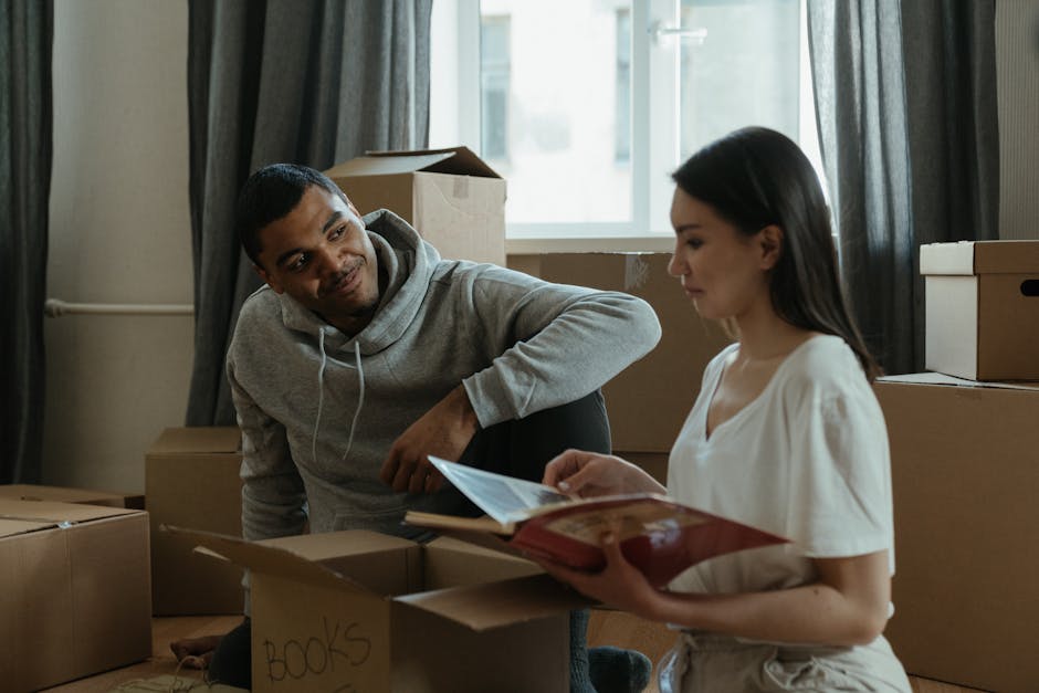 A man and a woman are inside a room surrounded by brown cardboard moving boxes, some labeled with black marker, including one with 'BOOKS'. The man, wearing a light grey hoodie, is kneeling on the floor, slightly leaning to his right with his right arm resting on his knee, and appears to be engaging in conversation. The woman, dressed in a white blouse, is holding open a folder or booklet and is looking at the man attentively. The room is lit by natural daylight coming through a window behind them, which is partially covered by dark curtains. The setting suggests a home relocation process involving packing or unpacking, with the presence of boxes and minimal furniture. The scene illustrates packing and moving activities, which is relevant to house removals and furniture transport services provided by Movers Balham, especially in relation to a home move like the SW12 Bedford Hill relocation described on the webpage.