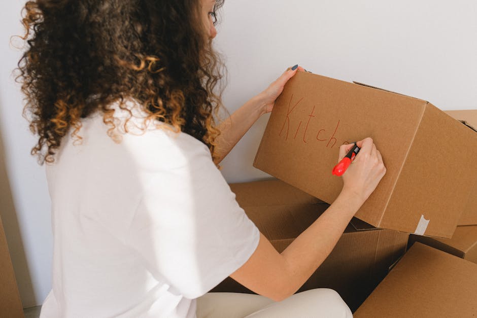 A woman with long, curly brown hair wearing a white shirt is inside a home, carefully writing the word 'kitchen' in red marker on a medium-sized cardboard box. She is seated on the floor surrounded by several other packed cardboard boxes, some of which are open and contain various household items. The scene takes place near a plain white wall, with natural light illuminating the area, indicating an ongoing home relocation or packing process. The woman appears to be cataloguing her belongings for a move, showcasing typical packing and moving activities that are part of house removals services. This image emphasizes the detailed preparation involved in furniture transport and packing for a house move, relevant to services offered by Movers Balham for SW12 renovations or Bedford Hill home moves, as described on their website.